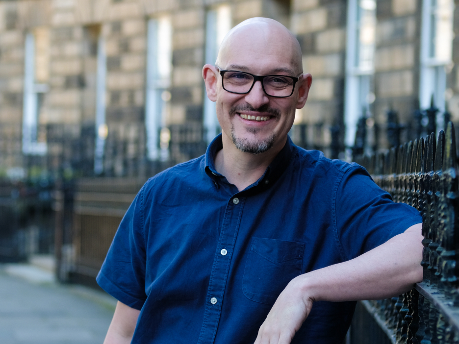 A photo of a broadly smiling white man in glasses wearing a dark blue short-sleeved shirt. He is leaning against a railing casually.